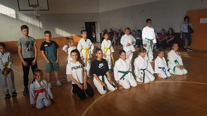 Grupa mladih karatista sa pojasevima različitih boja kleči i stoji u sportskoj sali dok roditelji posmatraju