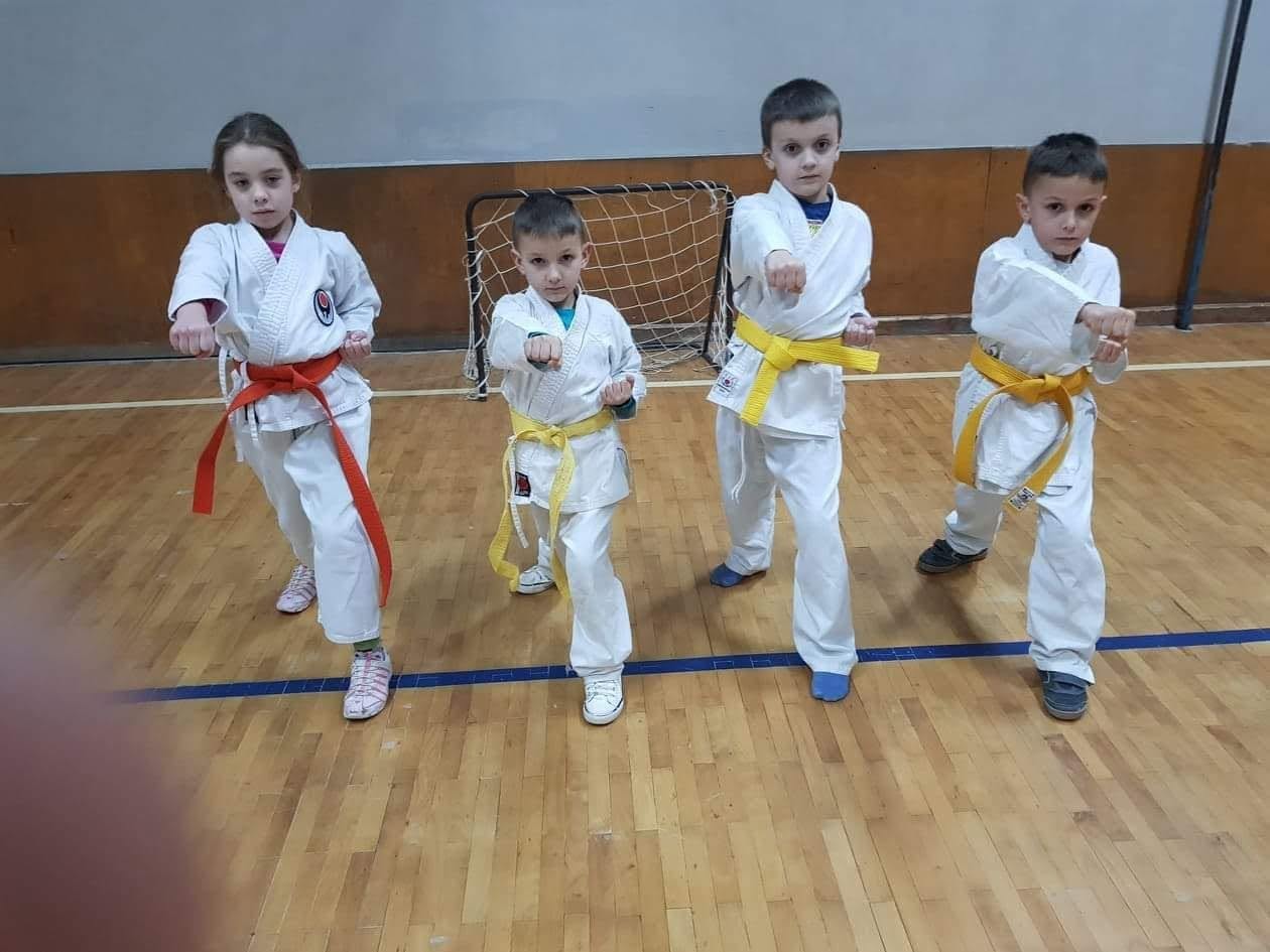 Četvoro djece u bijelim kimonima sa žutim i narandžastim pojasevima izvodi udarac u stavu, u sportskoj sali